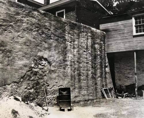 Black and white photo of a stone wall beside a building, with debris and tools scattered on the ground.