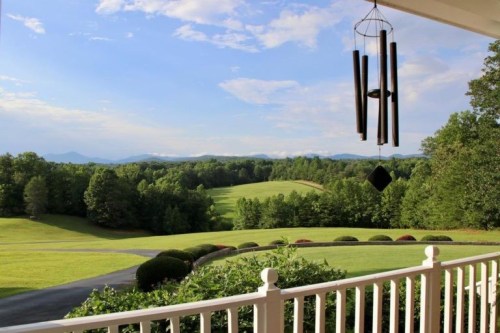 A scenic view from a porch overlooking lush green fields and distant mountains under a blue sky. Wind chimes hang nearby.