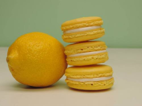 Three yellow macarons stacked next to a whole lemon on a light surface with a green background.