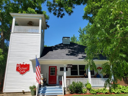 A charming white café with a red door and an American flag, surrounded by trees on a sunny day.