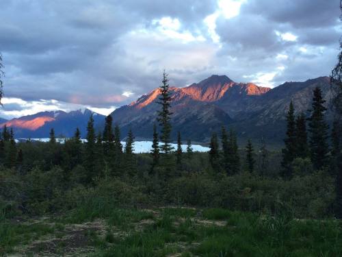 A scenic view of mountains with sunlight illuminating peaks, surrounded by trees and a lake under a cloudy sky.