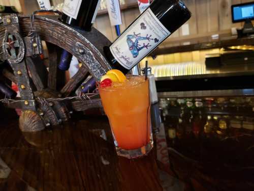 A colorful cocktail garnished with a slice of orange and a cherry, served on a wooden bar counter.