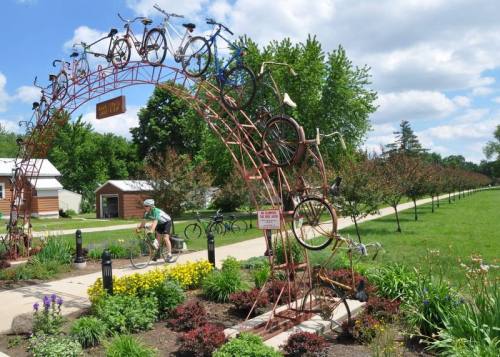 A bike-themed archway made of bicycles, with a cyclist passing by on a path surrounded by greenery and flowers.