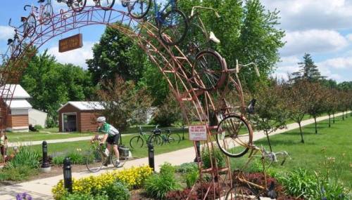 A cyclist rides under a decorative arch made of bicycles, surrounded by greenery and walking paths.