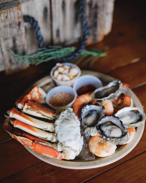 A platter of seafood featuring crab legs, shrimp, oysters, and small cups of sauces on a wooden table.