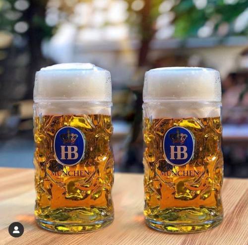 Two frosty beer mugs with foam, featuring the HB München logo, sit on a wooden table with a blurred background.