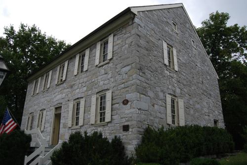 A historic stone building with white shutters, surrounded by greenery and featuring an American flag in front.