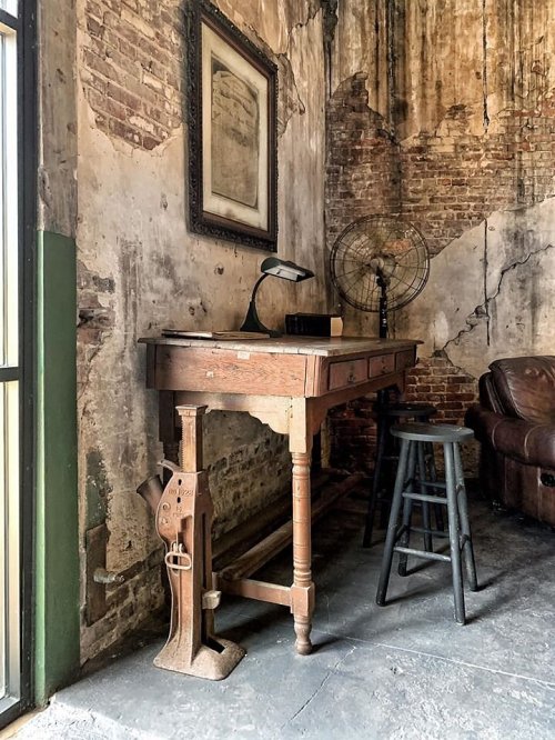 A rustic workspace featuring a wooden desk, a vintage fan, and a leather couch against a textured brick wall.