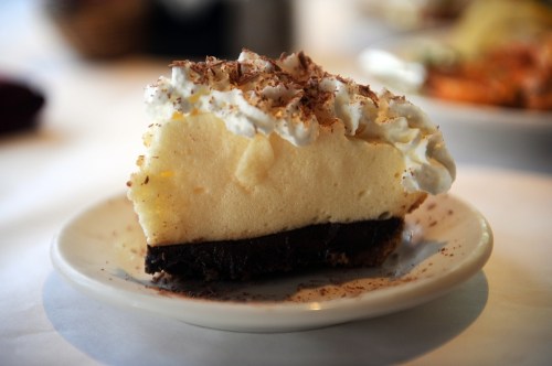 A slice of creamy cheesecake topped with whipped cream and chocolate shavings on a small plate.