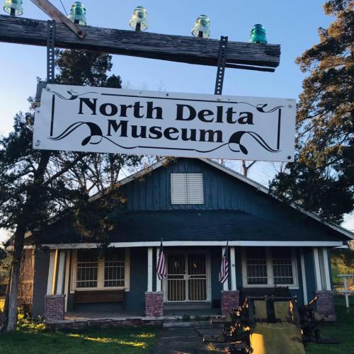 Sign for the North Delta Museum in front of a blue house, surrounded by trees and featuring American flags.