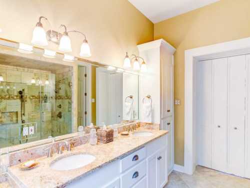 Bright bathroom with double sinks, granite countertop, large mirror, and glass shower. Light-colored walls and cabinetry.