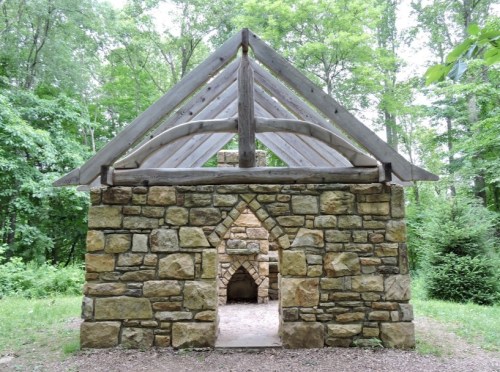 A stone structure with a wooden roof, set in a lush green forest. The entrance features an arched doorway.