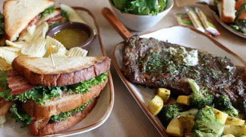 A table spread featuring a sandwich with lettuce and bacon, chips, a steak with herbs, and a side of vegetables.