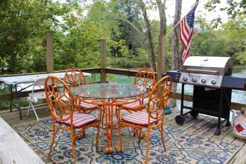 A cozy outdoor dining area with an orange table and chairs, a grill, and a view of greenery and water.