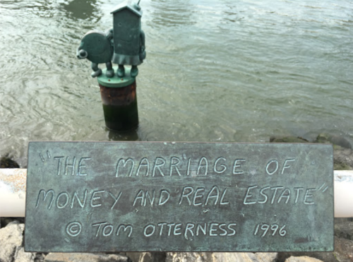 A plaque titled "The Marriage of Money and Real Estate" by Tom Otterness, beside a water feature.