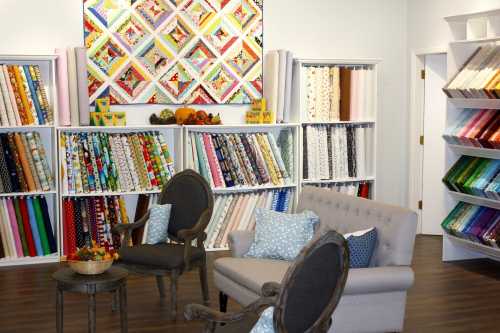 A cozy fabric store featuring colorful fabric rolls, a quilt on the wall, and a seating area with two chairs and a table.