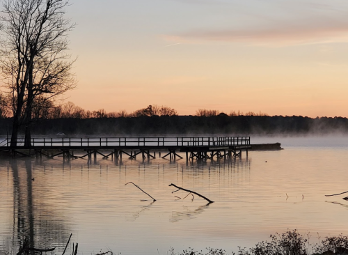 A serene lake at dawn with a wooden pier, mist rising from the water, and trees silhouetted against a colorful sky.