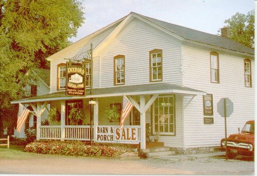 A white two-story building with a sign reading "Barn & Porch Sale," featuring American flags and flower planters.