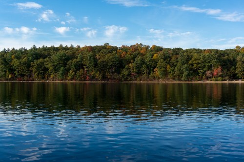 A serene lake reflecting a colorful tree line under a clear blue sky with scattered clouds.