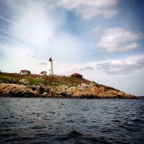 A lighthouse stands on a rocky shore, surrounded by green hills and a cloudy sky, with calm waters in the foreground.