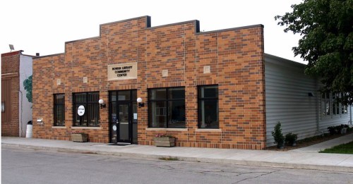 A brick building with a sign reading "Senior Library Community Center," featuring large windows and a welcoming entrance.