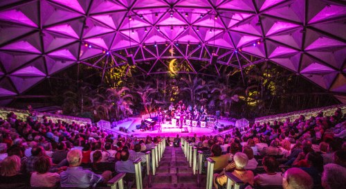 A vibrant concert scene with a large audience in a geometric outdoor venue, illuminated by purple lights.