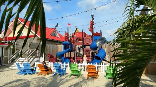 Colorful playground with slides and climbing structures, surrounded by palm trees and beach chairs on sandy ground.