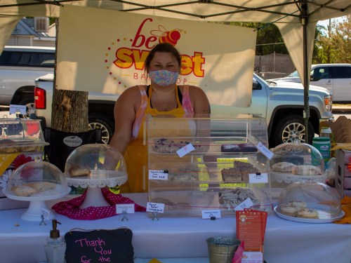 A baker stands behind a display of cakes and pastries under a tent, wearing a mask and apron, with a "Be Sweet" sign.
