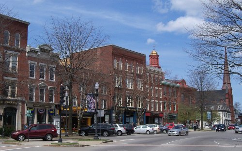 A charming downtown street with historic brick buildings, shops, and a church steeple under a blue sky.