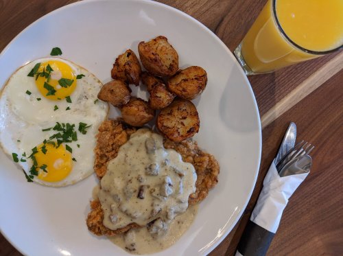 A plate of fried chicken with gravy, two sunny-side-up eggs, roasted potatoes, and a glass of orange juice.