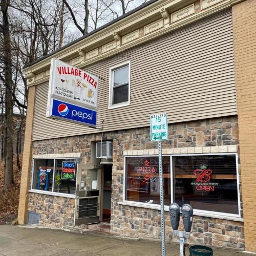 A pizza shop with a stone facade, featuring a sign for Village Pizza and nearby parking meters.