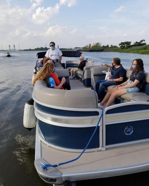 A group of people wearing masks relax on a pontoon boat on a calm river, with trees and buildings in the background.