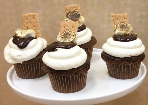 Four chocolate cupcakes topped with fluffy white frosting, chocolate drizzle, toasted marshmallows, and graham cracker pieces.