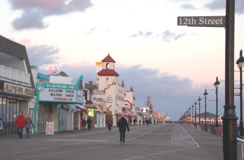 A boardwalk scene at sunset, featuring shops and a street sign for 12th Street, with people strolling along.