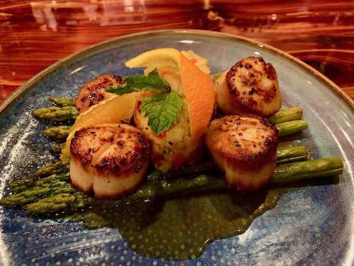 Seared scallops on a bed of asparagus, garnished with citrus and mint, served on a decorative blue plate.