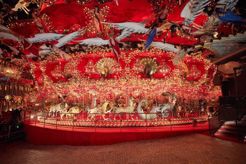 A vibrant carousel adorned with lights and decorations, set against a red backdrop with whimsical figures overhead.