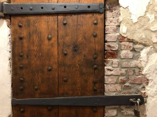 A weathered wooden door with metal reinforcements and a padlock, set against a brick wall with peeling paint.