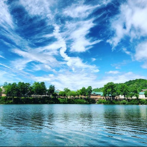 A serene lake scene with clear blue skies, fluffy clouds, and lush greenery along the shoreline.