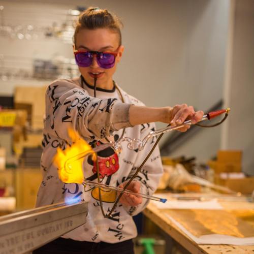 A person wearing sunglasses and a Mickey Mouse sweater is using a torch to create flames in a workshop setting.