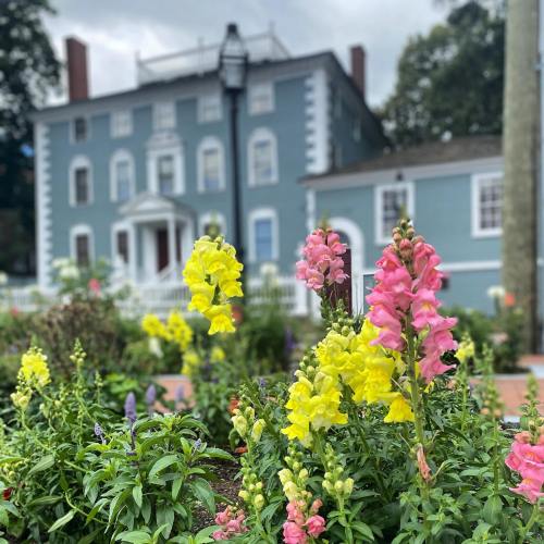 A historic blue house with a garden of colorful flowers in the foreground, under a cloudy sky.