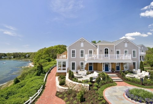 A beautiful waterfront house with a spacious deck, surrounded by greenery and a winding brick pathway.