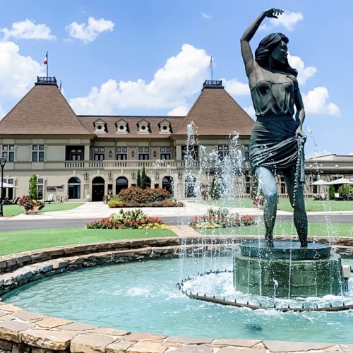 A statue of a woman stands in a fountain, with a historic building and blue sky in the background.