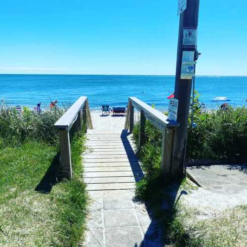 A wooden walkway leads to a beach with calm blue waters and sunbathers in the distance.