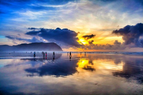 Silhouettes of people walking on a wet beach at sunset, with vibrant clouds and reflections in the water.