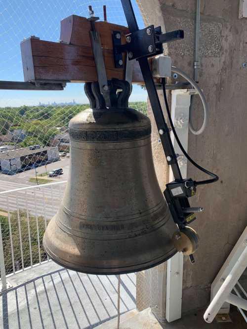 A large metal bell hanging from a mechanism, with a view of a landscape in the background.