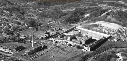 Aerial view of a large industrial complex surrounded by hills and residential areas, in black and white.