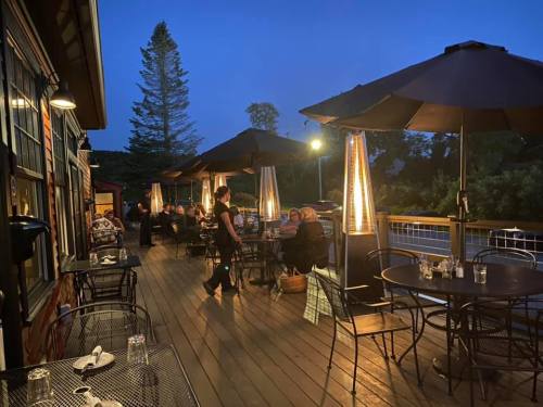 Outdoor dining area at dusk, featuring tables with umbrellas and patrons enjoying their meals. Warm lighting creates a cozy atmosphere.