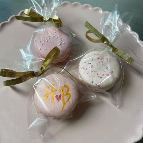 Three decorated macarons in clear bags with gold ribbons, featuring pink and white colors and festive designs.