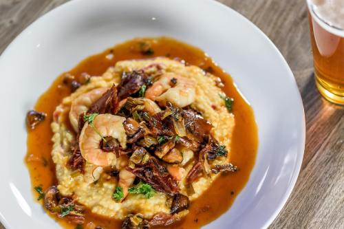 A plate of creamy grits topped with shrimp, sautéed vegetables, and a rich sauce, alongside a glass of beer.