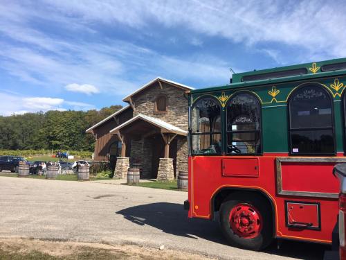 A colorful trolley bus parked near a rustic stone building surrounded by trees and barrels.